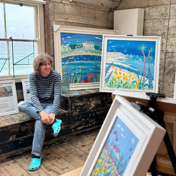 Cornish artist John Dyer pictured at Porthmeor Studios in St Ives, seated by the window and surrounded by his vibrant Cornish paintings.