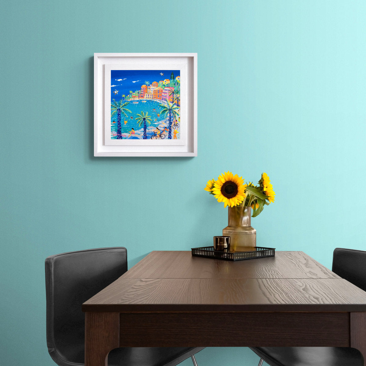 Framed artwork on a teal wall above a wooden table with sunflowers. 'Cycling by the Bay, Villefranche-sur-Mer'. 12 x 12 inches acrylic on canvas. Painting of the South of France by John Dyer