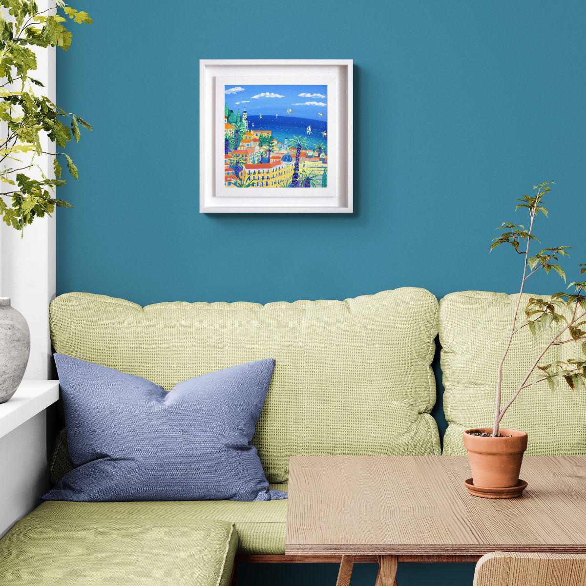 Green sofa with blue pillow, framed artwork on blue wall, and potted plant. 'View from the Winter Palace, Menton'. 12 x 12 inches acrylic on canvas. Painting of the South of France by John Dyer