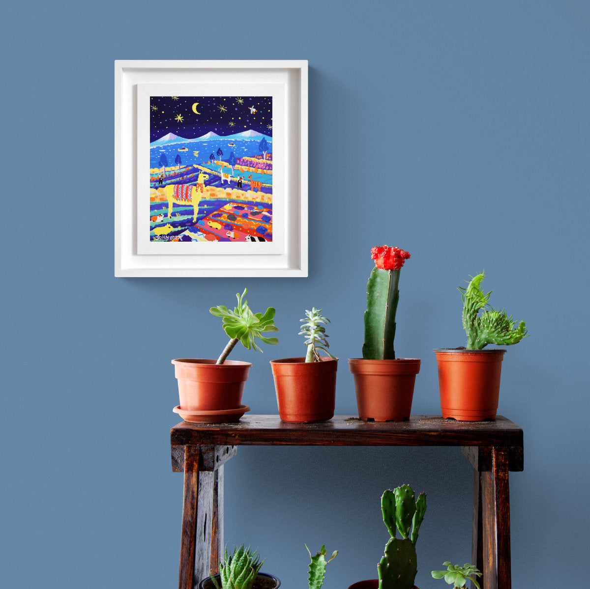 Framed artwork above a wooden shelf with potted plants against a blue wall. Colorful artwork of llamas in a starry night landscape with mountains and people. 'Moonlit Llamas and Guinea Pigs at the Potato Harvest, Peru’, 12x10 inches acrylic on canvas. Painting of Peru by British Artist John Dyer