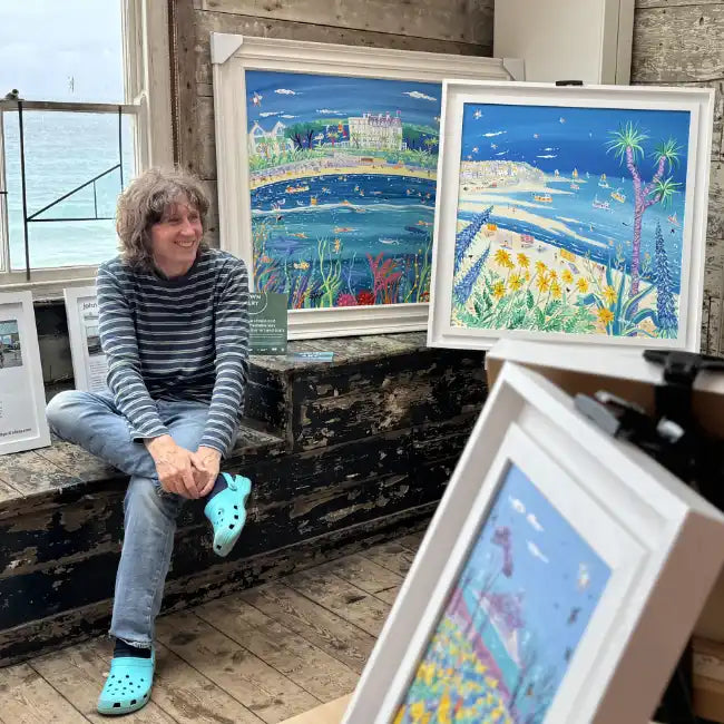 Cornish artist John Dyer sitting with framed artworks depicting coastal scenes in Porthmeor Studios St Ives, Cornwall