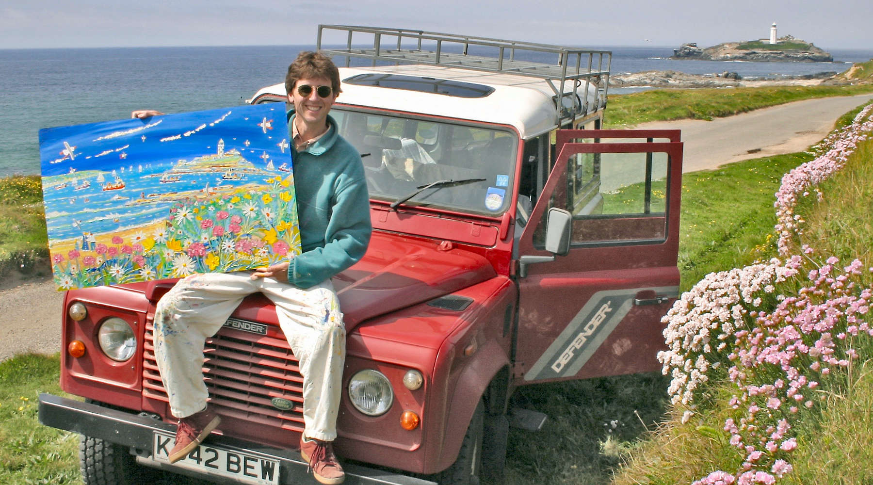 Cornish artist John Dyer sitting on his Land Rover 110 Defender with a painting of Godrevy lighthouse in Cornwall.