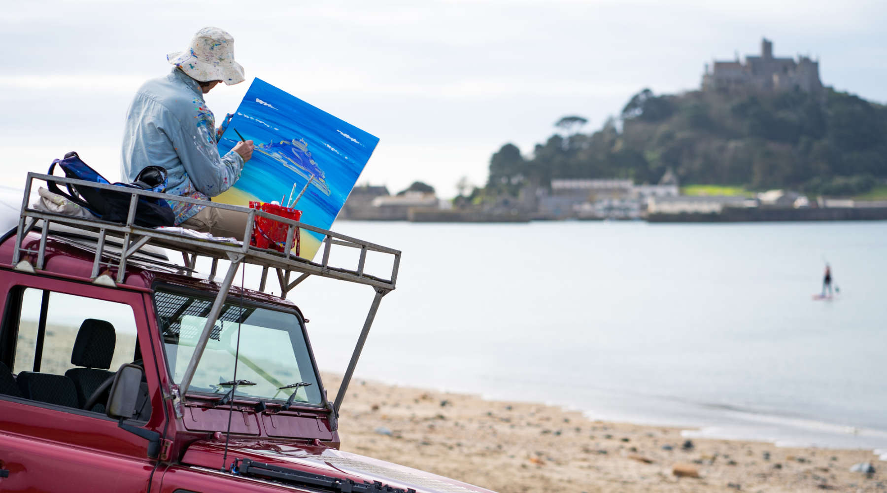 Artist John Dyer painting St Michael's Mount on his Land Rover Defender 110 at Marazion beach in Cornwall