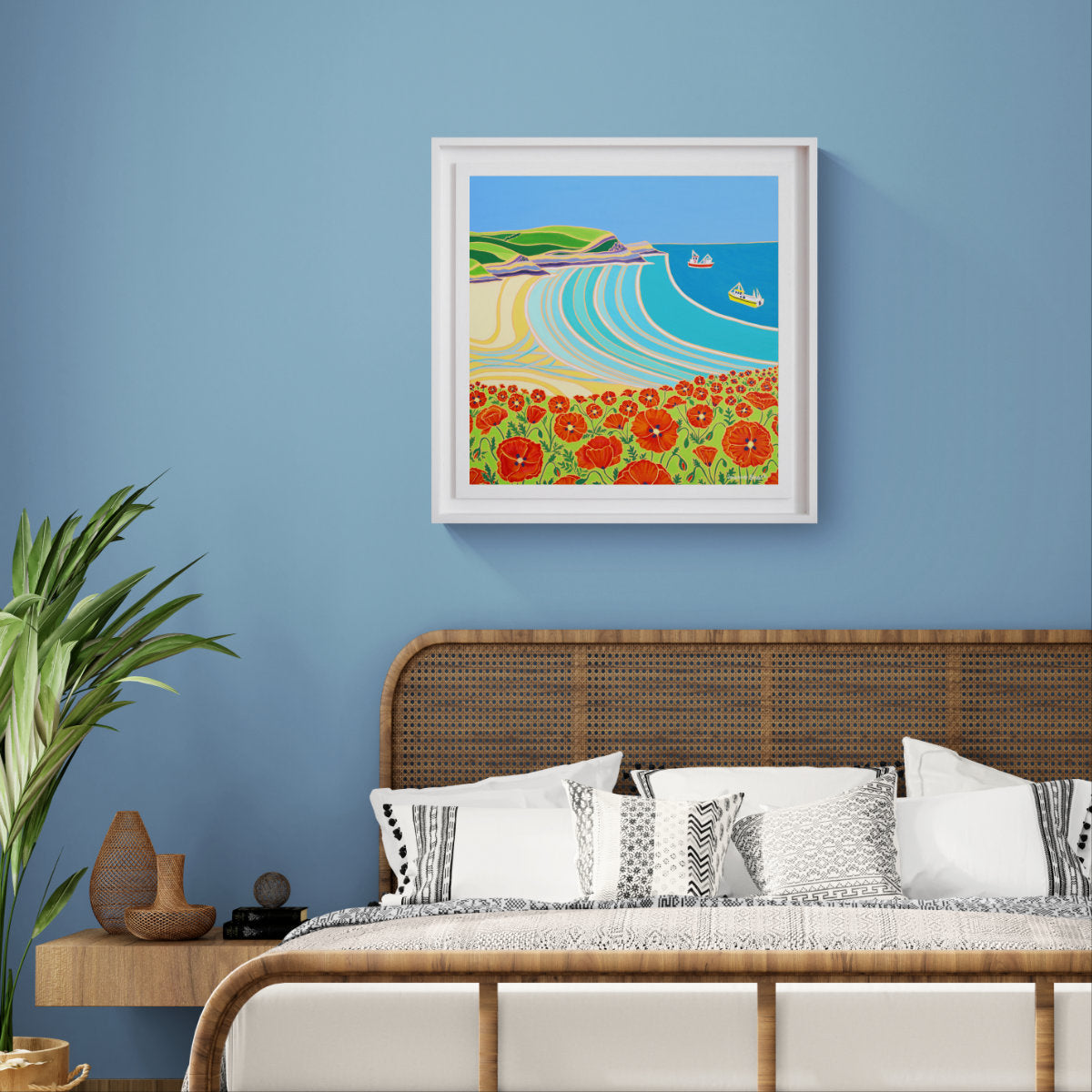 A framed original oil painting depicting a coastal landscape with red poppy flowers, green hills, and blue waves, displayed on a light blue wall above a bed with white and patterned pillows. Joanne Short Original Oil Painting of Cornwall. 'Poppies Galore, Polly Joke'. 24 x 24 inches oil on canvas.
