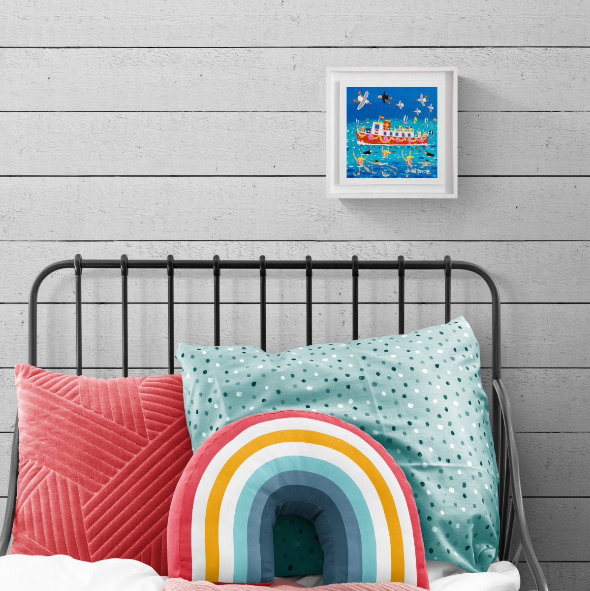 A colourful framed painting featuring a Cornish ferry boat and seagulls styled on a light grey wood-paneled wall above a bed with pink and mint pillows and a rainbow cushion. 'Ferry Time Fun and Skinny Dippers, Falmouth’, 8 x 8 inches acrylic on canvas. Painting of Cornwall by Cornish Artist John Dyer