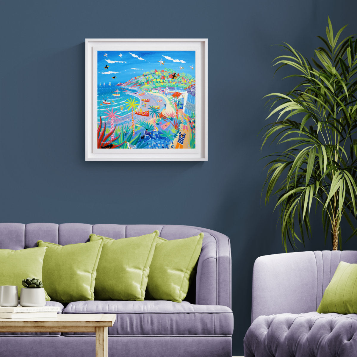 A colourful framed original acrylic painting depicting a vibrant sub-tropical Cornish scene, displayed on a dark blue wall above a purple sofa in a modern living room setting. 'Coastal Walk, Porthallow', 24 x 24 inches acrylic on canvas. Original Painting by Cornish Artist John Dyer.