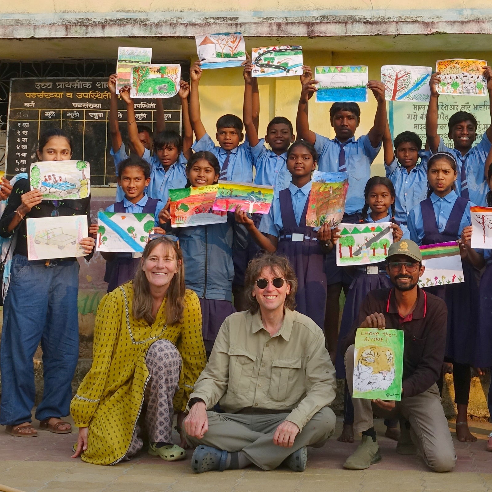 Group of Gond children in India holding up their Last Chance to Paint artworks with artists Joanne Short and John Dyer posing for a photo in front of a chalkboard.