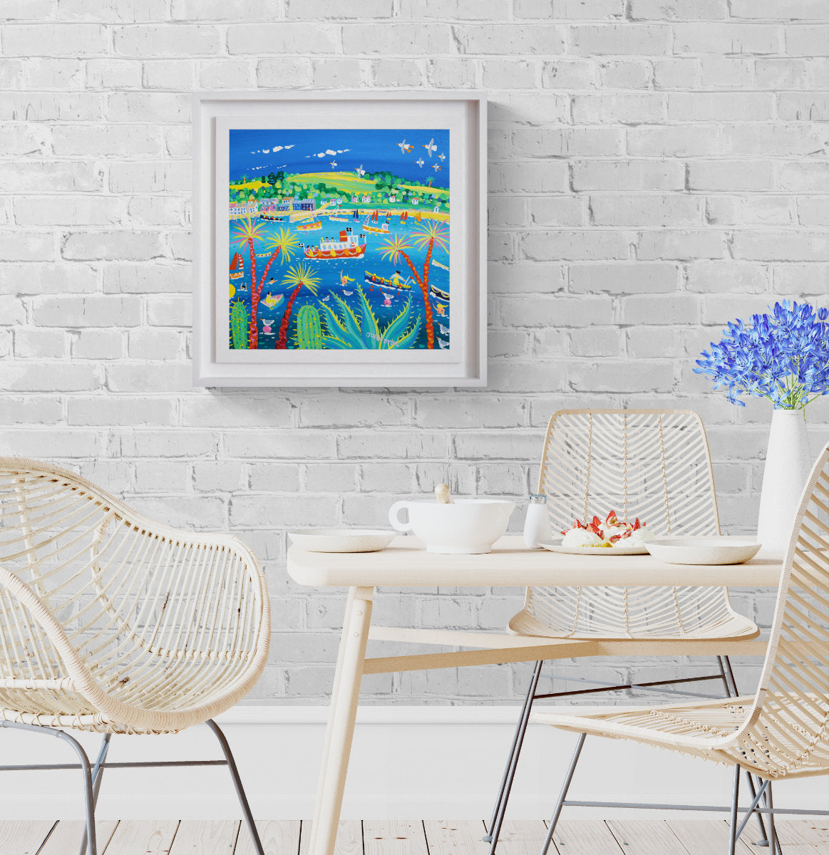 A colourful framed Cornish painting depicting a tropical harbour scene with boats, palm trees, and blue sky, displayed on a white brick wall above a dining table with woven chairs. 'Summer Colours on the River, Falmouth’, 18 x 18 inches acrylic on canvas. Cornish Coastal Art. Painting by Artist John Dyer