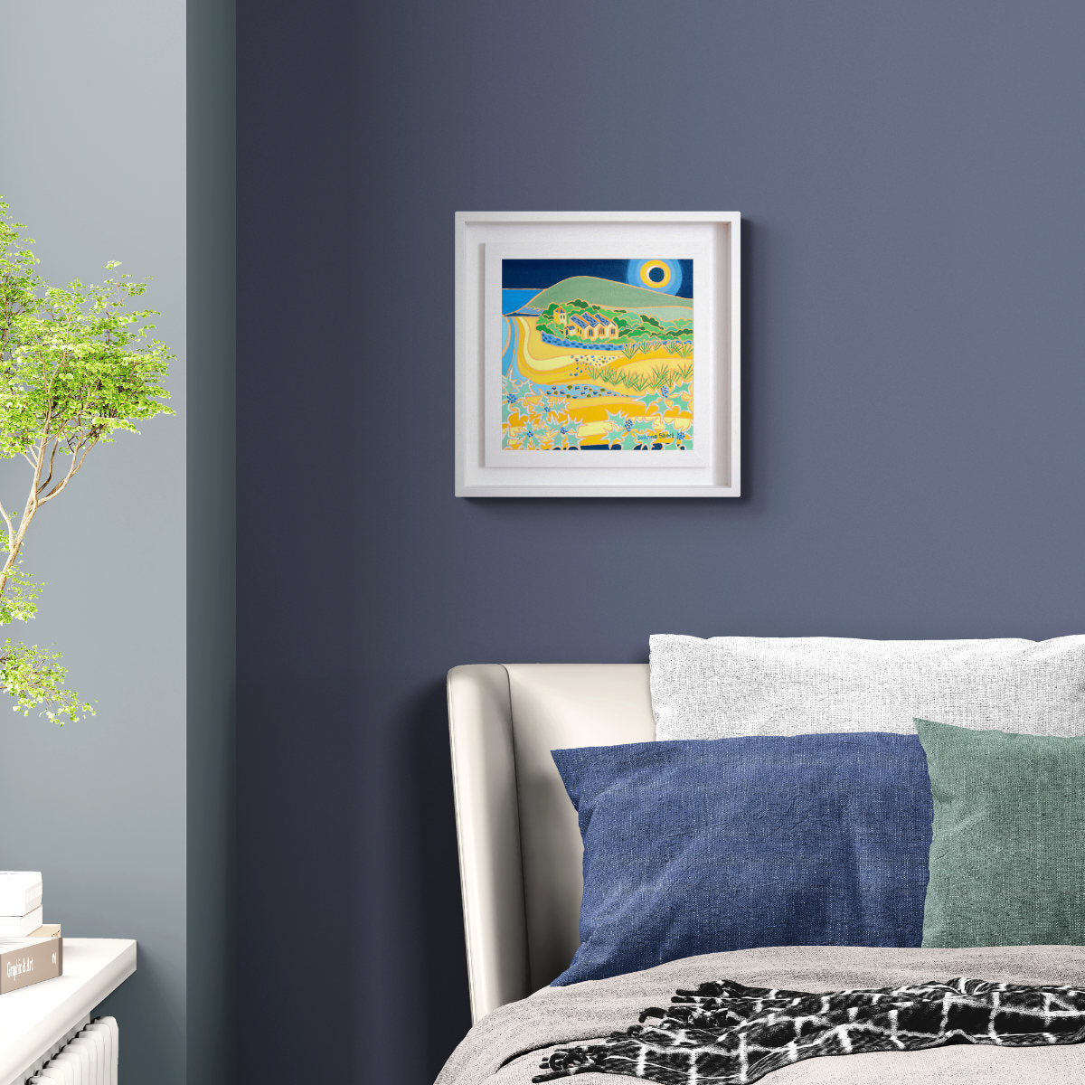 A framed colorful landscape art print hangs on a dark blue-gray wall above a bed with blue and green pillows in a modern bedroom setting. 'Eclipse over the Church, Gunwalloe Church Cove'. 12 x 12 inches Original Oil Painting on Canvas by Cornish Artist Joanne Short