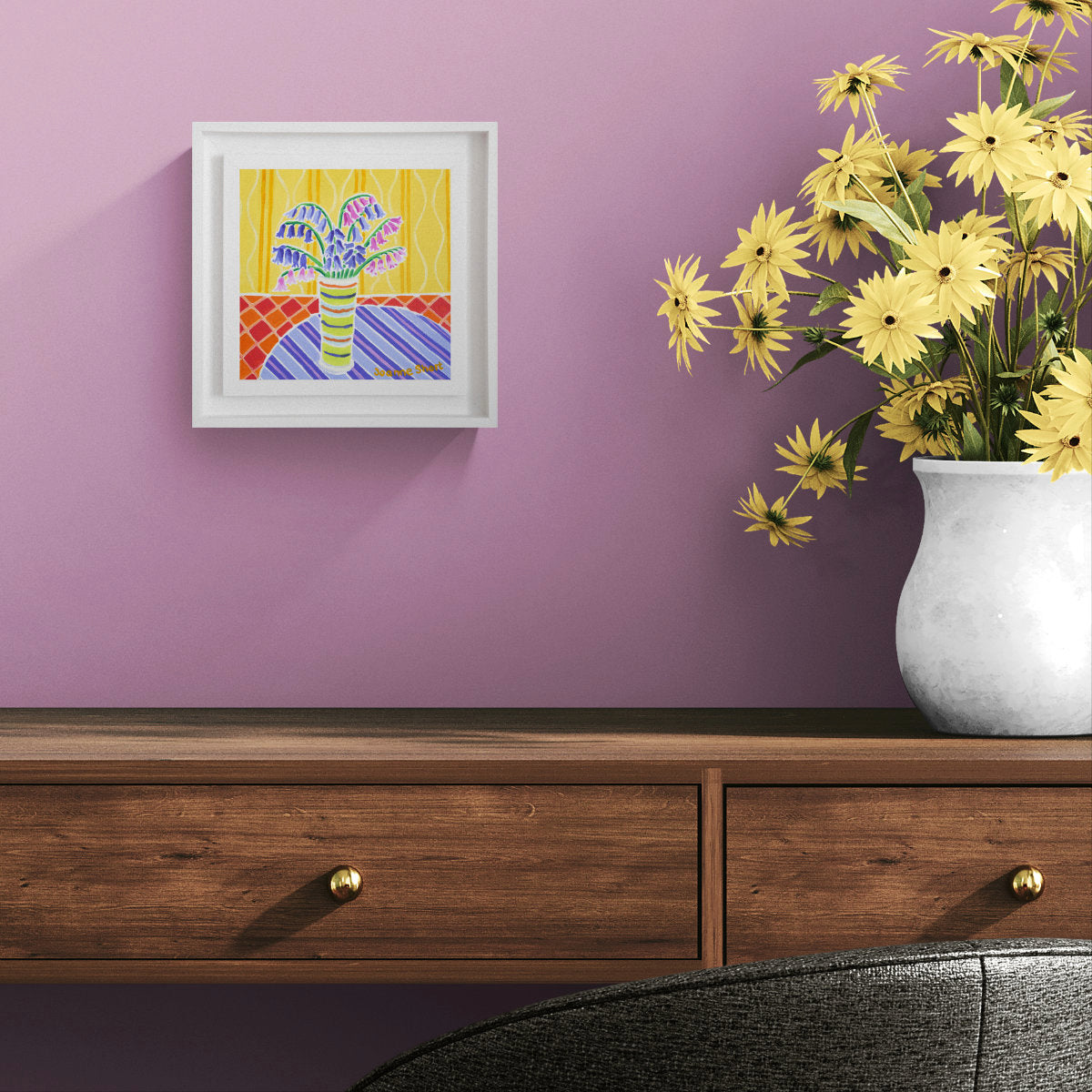 A framed colourful oil painting depicting a striped vase with flowers, displayed on a lavender wall above a wooden console table with a white vase of yellow flowers. 'Flowers from a Friends Garden', 8 x 8 inches oil on canvas flower still life art. Painting by Cornish Artist Joanne Short