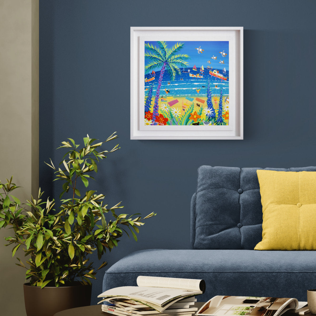 A framed colourful beach painting hanging on a dark blue wall above a blue sofa with a yellow pillow, next to a potted plant and stacked magazines. 'Hot Summer Day, Gyllyngvase Beach, Falmouth’, 18 x 18 inches acrylic on canvas. Original Painting by Cornish Artist John Dyer
