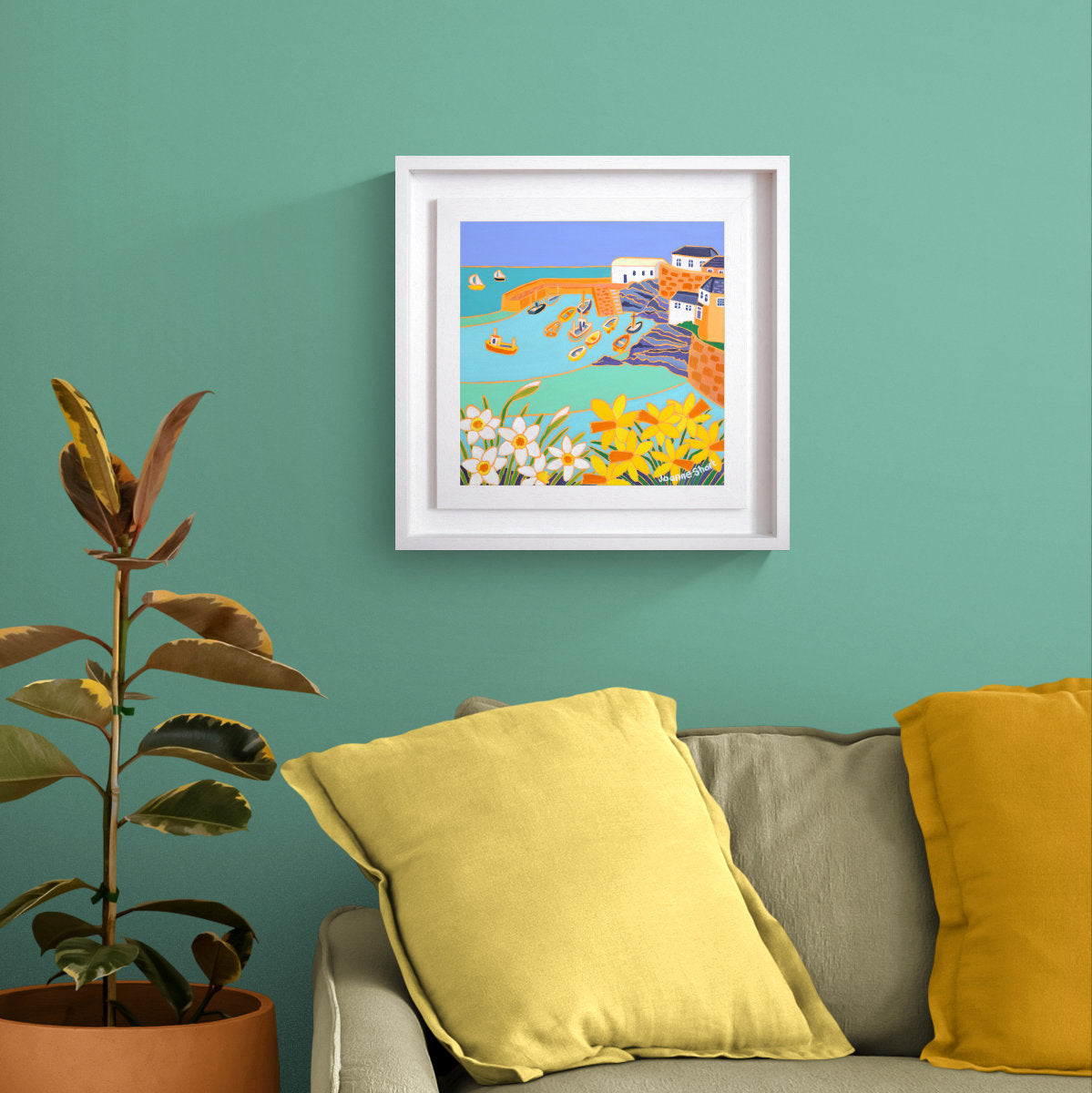 A framed oil painting depicting a colourful coastal village scene with flowers, hung on a teal wall above a sofa with yellow pillows and a potted plant. 'Spring Tide, Coverack'. 12 x 12 inches Original Oil Painting on Canvas by Cornish Artist Joanne Short