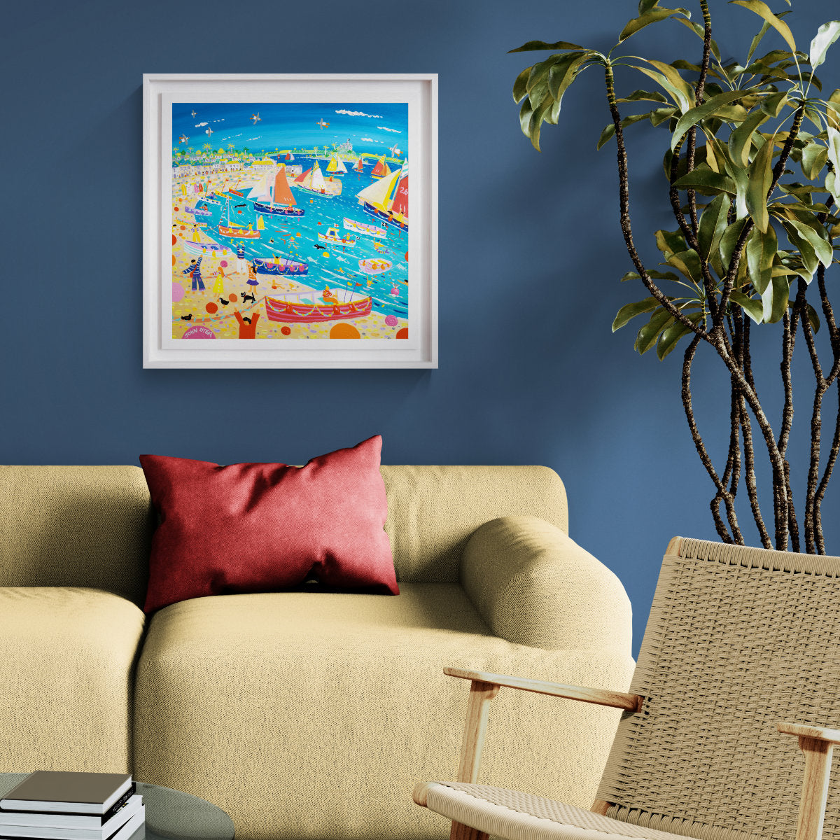 A colourful framed art print depicting a vibrant beach and harbour scene, hung on a dark blue wall above a beige sofa with a red pillow in a styled living room. 'Sea Salts and Sail Festival, Mousehole', 24 x 24 inches acrylic on canvas. Original Painting by Cornish Artist John Dyer.