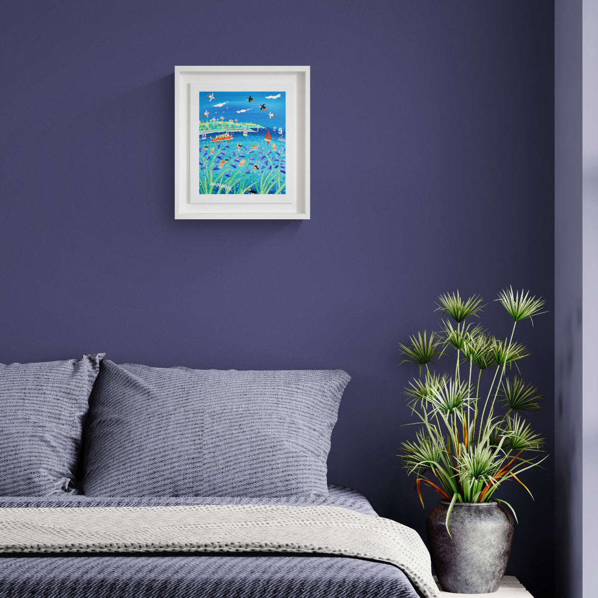 A framed original painting depicting a colourful landscape with blue sky, green foliage, and small boats, displayed on a dark purple wall above a bed with grey pillows and a potted plant nearby. 'Bathing Bluebelles , Falmouth', 12 x10 inches acrylic on canvas by Cornish Artist John Dyer
