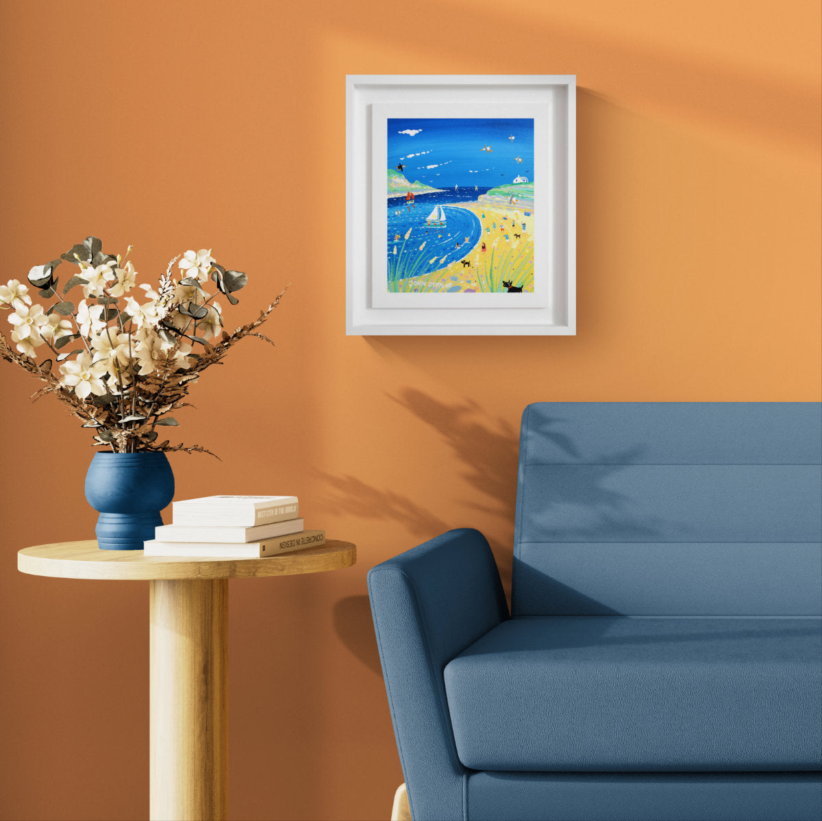 A framed original painting depicting a vibrant blue coastal landscape with sailboats and a sandy beach, styled on an orange wall above a blue sofa and next to a wooden side table with a blue vase of dried flowers. 'Day on the Beach, Daymer Bay', 12 x10 inches acrylic on canvas by Cornish Artist John Dyer