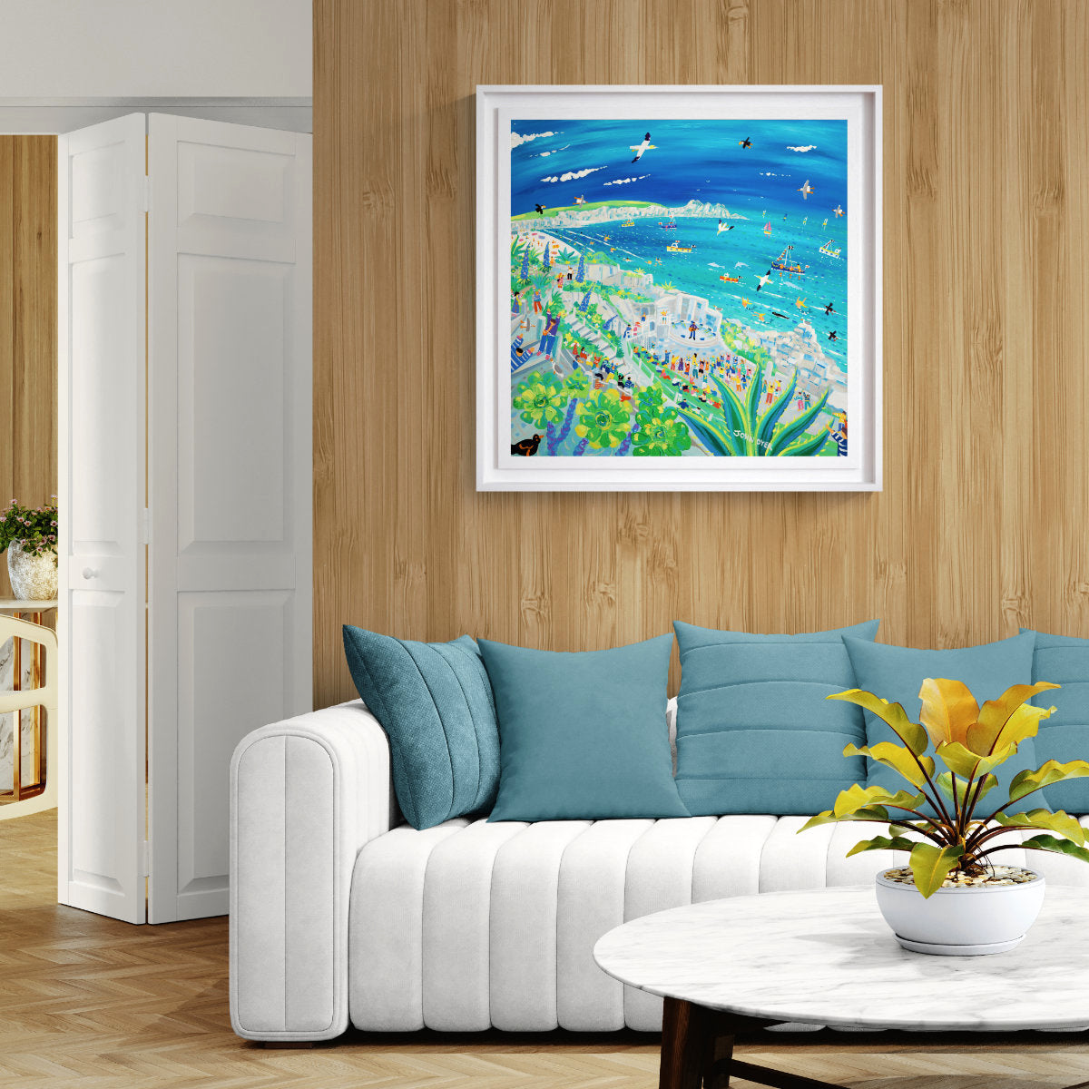 A framed colourful beach painting hangs on a light wood wall above a white sofa with blue pillows in a modern living room setting. 'Song on the Cornish Breeze at the Minack Theatre', 30 x 30 inches acrylic on canvas. Cornish Coastal painting by Cornish Artist John Dyer