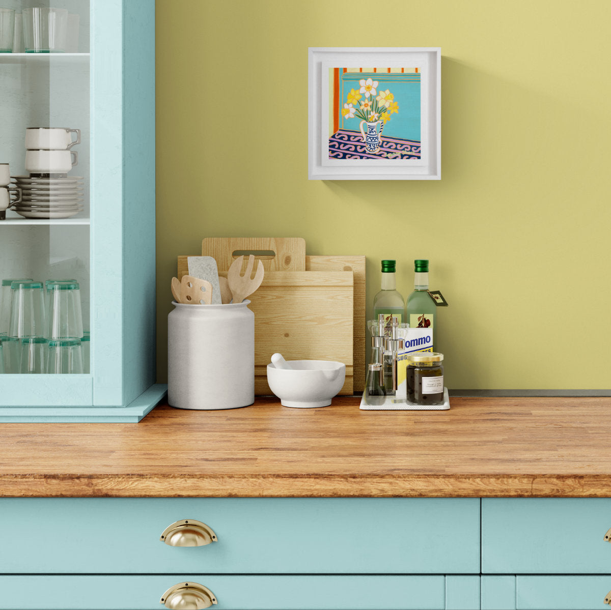 A framed original oil painting of a colourful floral table scene hangs on a light olive-green wall above a wooden countertop in a kitchen setting. The kitchen has light blue cabinets, a white ceramic jar with wooden utensils, cutting boards, and bottles. 'Cornish Daffodils in a Vase', 8 x 8 inches oil on canvas flower still life art. Painting by Cornish Artist Joanne Short