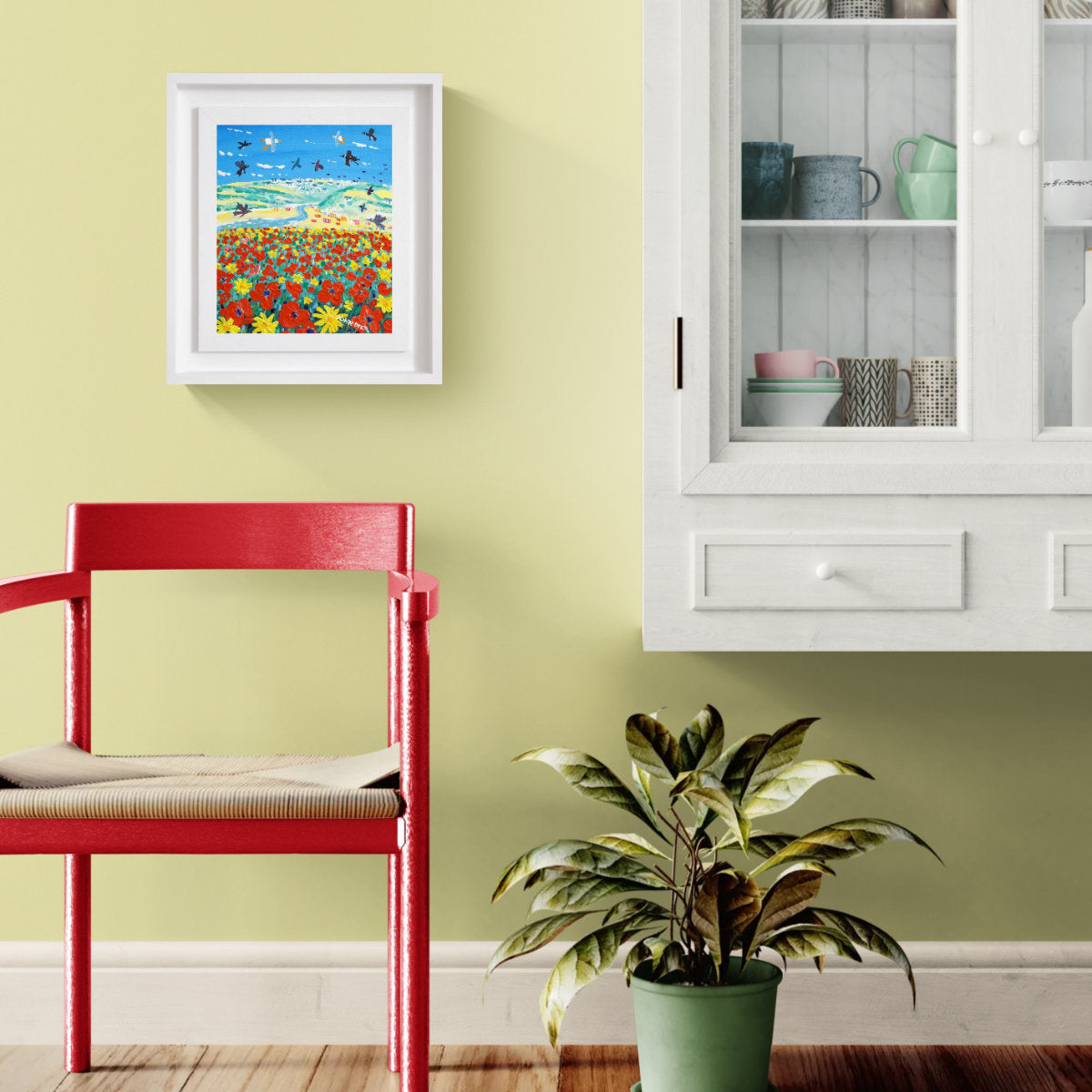A framed colourful art print depicting a field of red and yellow flowers under a blue sky with birds, hung on a light green wall in a styled room with a red chair and a potted plant. 'Skylarks and Poppies, Crantock Beach', 12 x10 inches acrylic on canvas by Cornish Artist John Dyer
