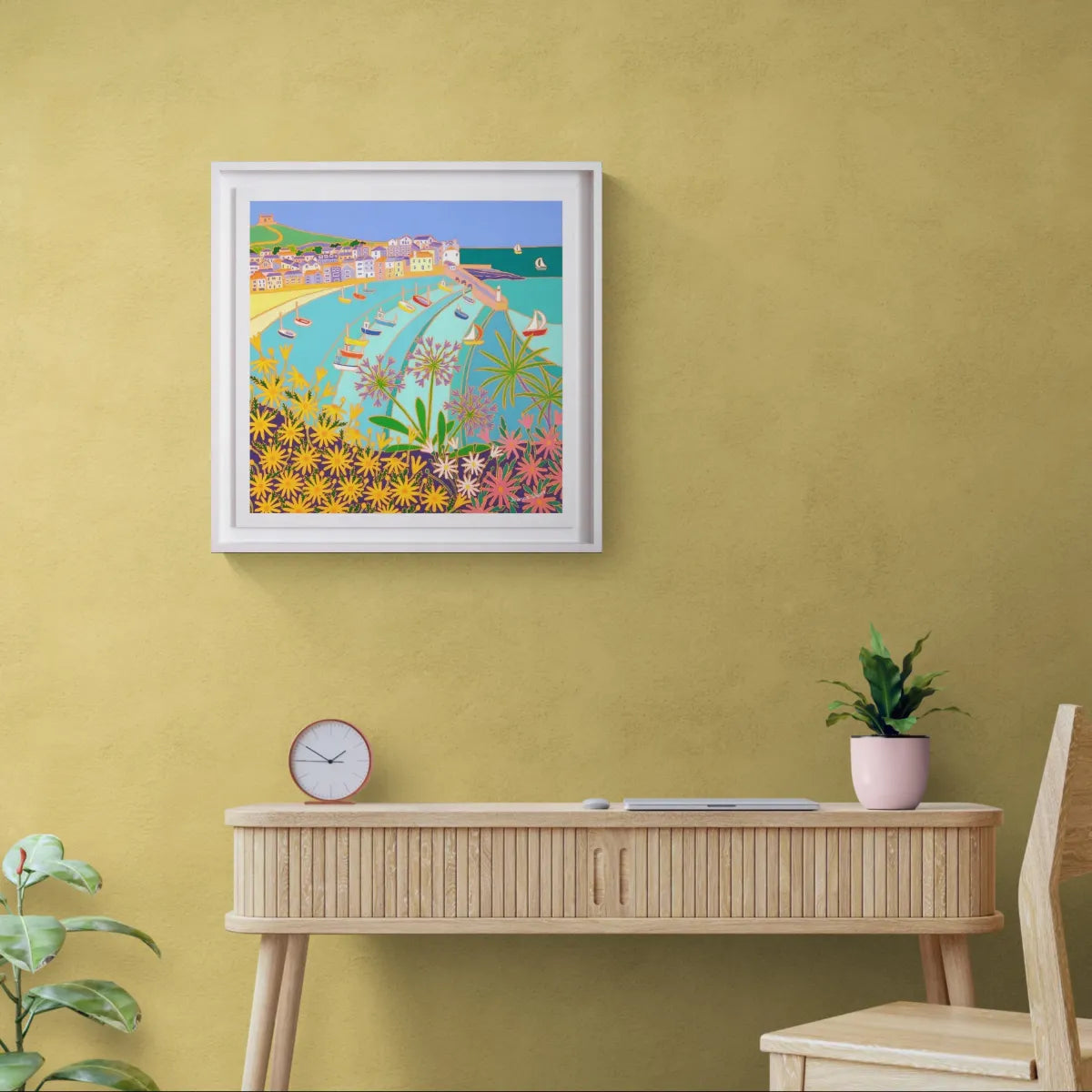 Framed artwork on a yellow wall above a wooden console table with a plant and clock. 'Sunny Seaside Flowers, St Ives’. 24x24 inches oil on canvas. Original Garden Painting of Cornwall by Cornish Artist Joanne Short.