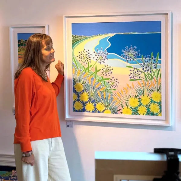 Cornish artist Joanne Short standing next to a colourful framed original painting of a Tresco beach scene with flowers.