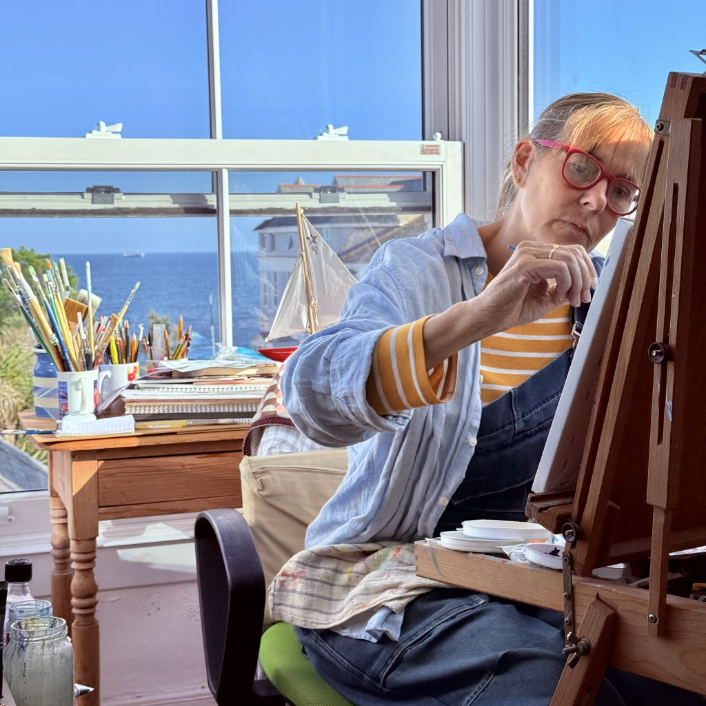 Cornish artist Joanne Short is painting at her easel, with a scenic view of the sea and sky through her studio windows in Falmouth. Paintbrushes and a model boat are on her desk, along with many drawing books.
