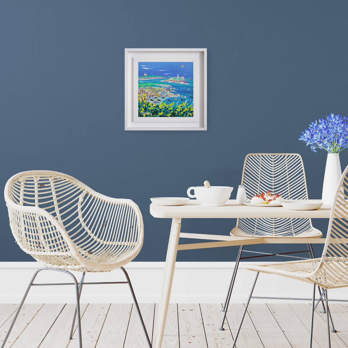 Modern dining area with wicker chairs, a table, and a framed artwork on a blue wall with 'Golden Gorse and Cornish Seals, Godrevy Lighthouse', 12x12 inches acrylic on canvas. Original Painting by Cornish Artist John Dyer.