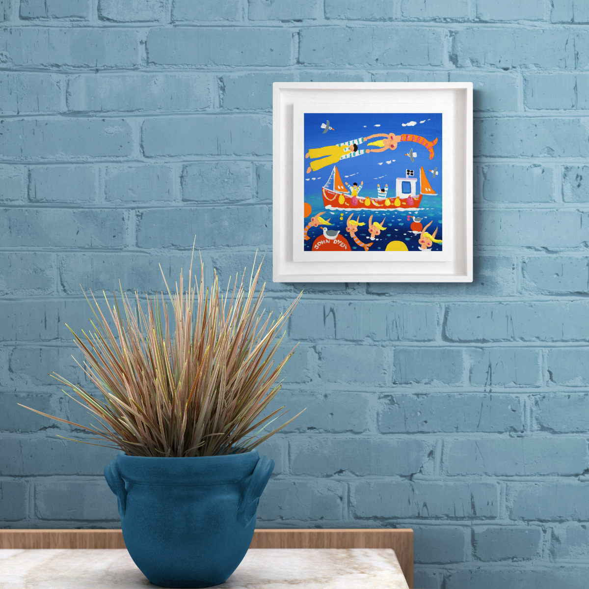 Framed artwork of a colorful scene with a boat and fish on a blue brick wall, below a potted plant. 'Cornish Mermaids’, 8x8 inches acrylic on canvas. Painting of Cornwall by Cornish Artist John Dyer