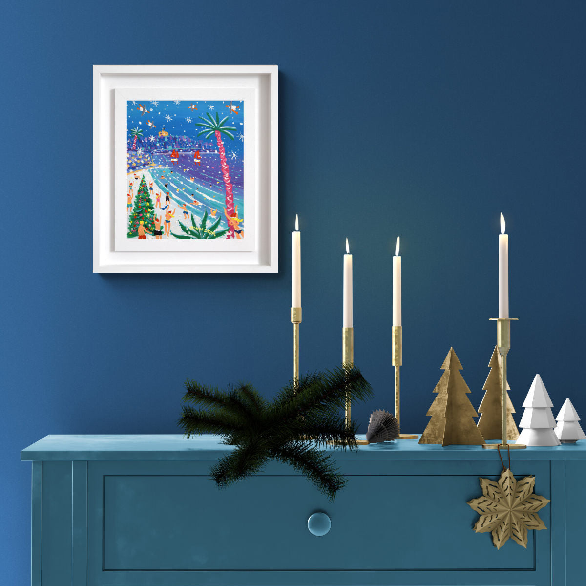 Decorative setup with framed artwork, candles, and Christmas trees on a blue surface against a blue wall. 'Gylly Beach Christmas Swimmers’, 12x10 inches acrylic on canvas. Painting of Cornwall by Cornish Artist John Dyer