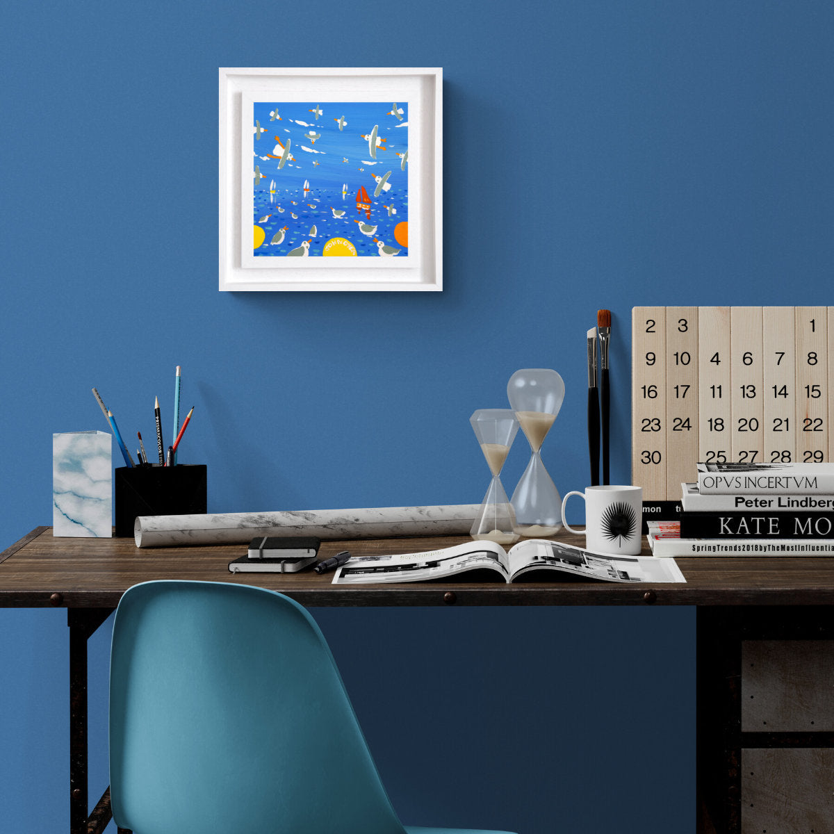 Desk with office supplies against a blue wall with a framed picture. 'Seagulls and Sailing Boats, Cornwall’, 8x8 inches acrylic on canvas. Painting of Cornwall by Cornish Artist John Dyer