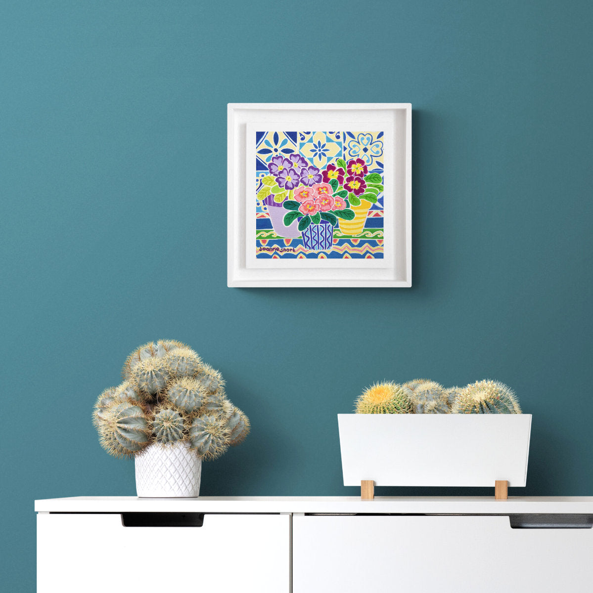 Framed artwork with floral design above a white cabinet with decorative plants on a teal wall. 'Spring Flowers on a Patterned Tablecloth', 8 x 8 inches oil on canvas still life. Painting by Cornish Artist Joanne Short