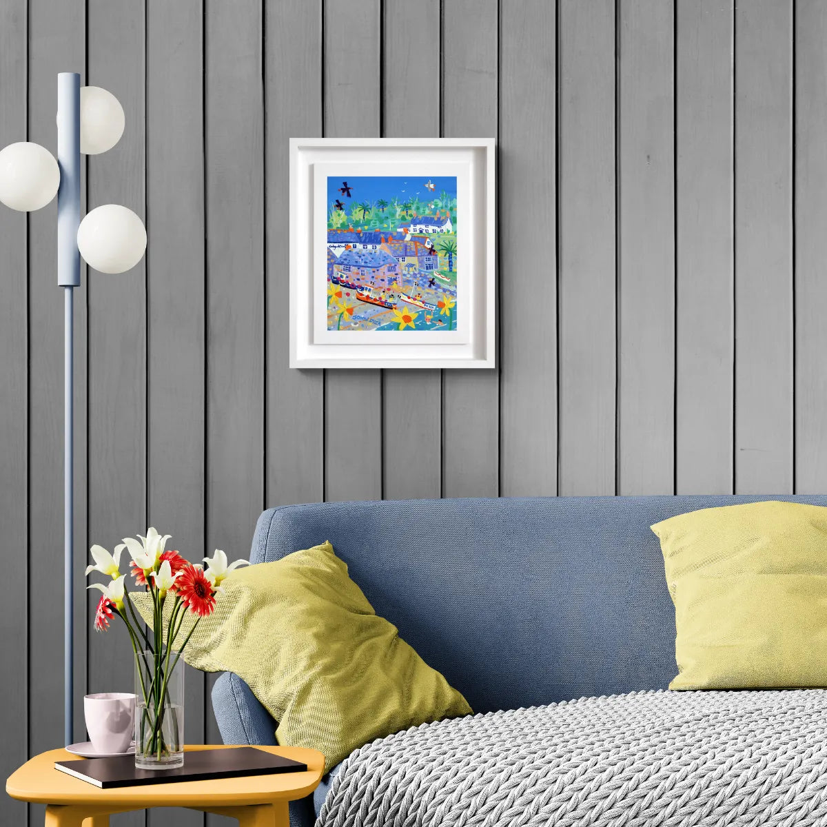 Living room with a blue sofa, yellow pillows, a framed painting on the wall, and a side table with flowers. 'Daffodils, Choughs and Boats, Cadgwith Cove', 12x10 inches acrylic on canvas. Painting of Cornwall by Cornish Artist John Dyer