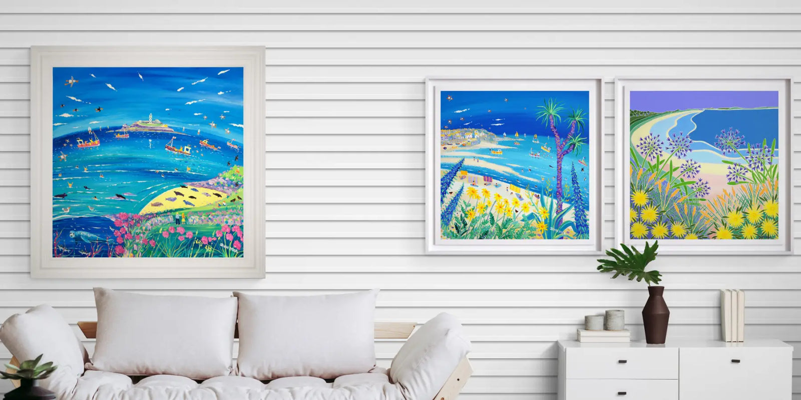 Three framed original painting artworks depicting coastal scenes with boats and landscapes on a white wall. Artists John Dyer and Joanne Short. Coastal interior setting.