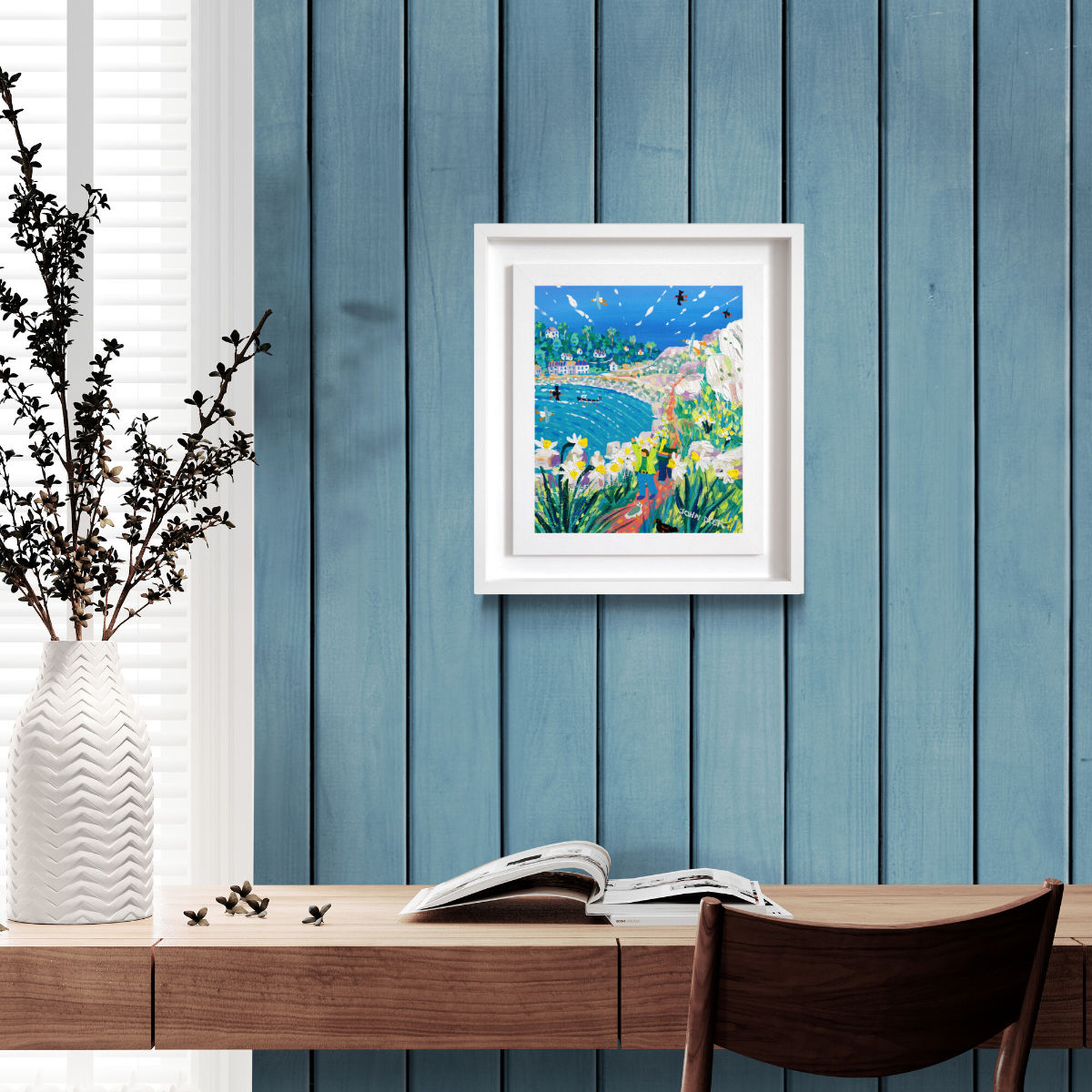 Framed artwork on a blue wall above a wooden desk with a vase and chair. 'Spring Walk to Lamorna Cove', 12x10 inches acrylic on canvas. Painting of Cornwall by Cornish Artist John Dyer