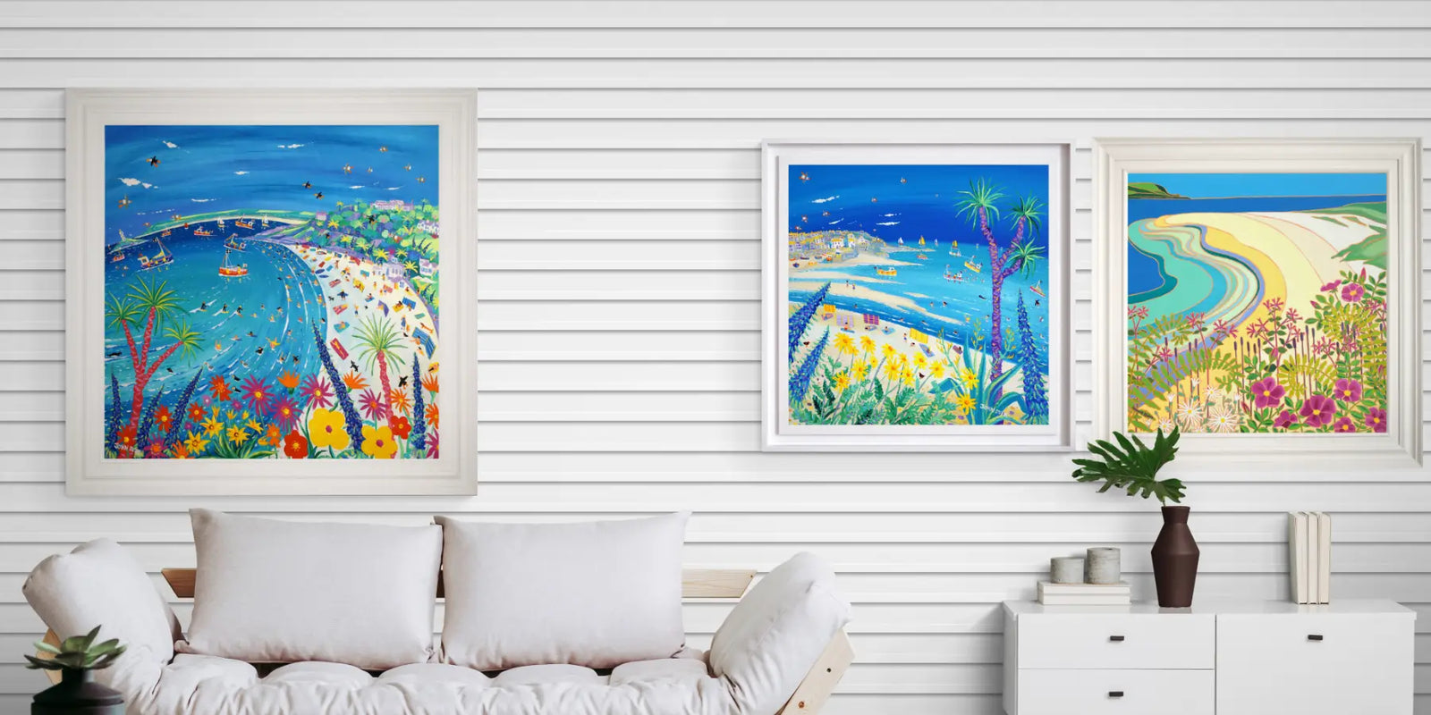 Three colourful framed paintings of coastal scenes on a white wall above a sofa. Original paintings of Cornwall by John Dyer and Joanne Short