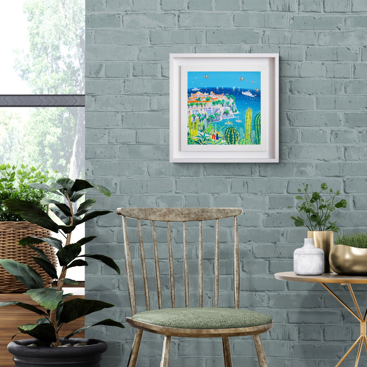 Framed artwork on a gray brick wall with a chair and plants in the foreground. 'Exploring the Jardin Exotique, Monaco'. 12 x 12 inches acrylic on canvas. Painting of Monaco, France by John Dyer