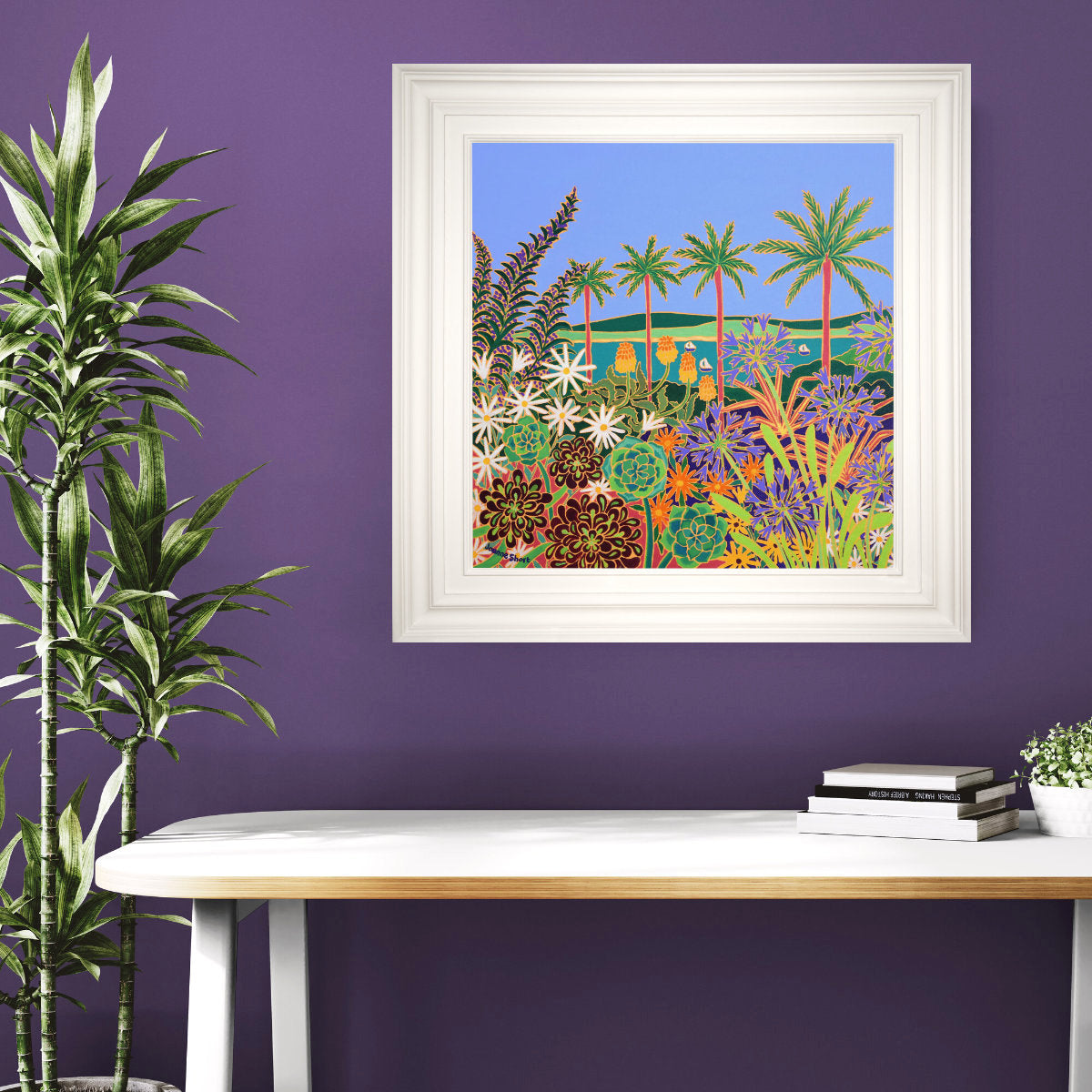 Framed tropical landscape painting on a purple wall with a white desk and plant. 'Botanical Symphony, Tresco Abbey Gardens’. 24x24 inches oil on canvas. Painting of Cornwall by Cornish Artist Joanne Short