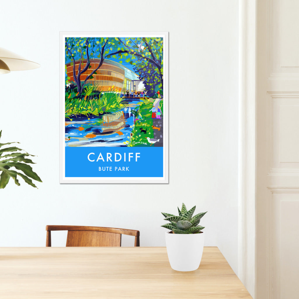 This stunning wall art travel poster print by John Dyer features the Royal Welsh College of Music and Drama in Bute Park, Cardiff in Wales. A family stroll through the park under the trees alongside the river. A duck, seagulls, scotty dog and sausage dog look on and on the bridge to the RWCMD musicians wave their musical instruments in the air celebrating their musical lives. Available unframed or framed and ready to hang in your home, student digs, business or office.
